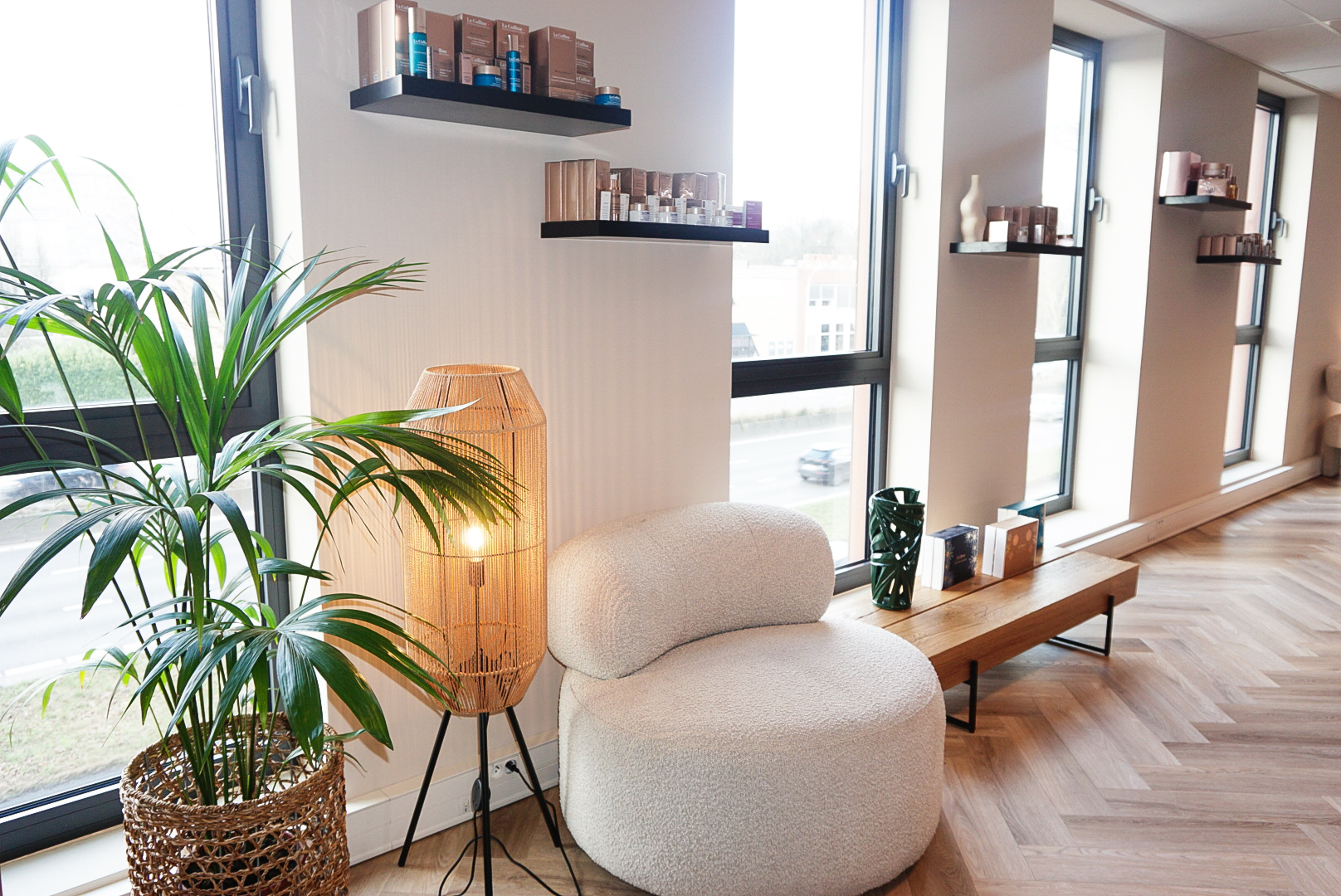 Lichte ruimte in MED-CLINIC Antwerpen met wandrekken vol huidverzorgingsproducten, een crèmekleurige fauteuil, rieten staanlamp en een palmplant bij de ramen.